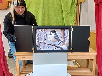 Marc-Norman Künnecke liest im Fröbel-Kindergarten und Familienzentrum Kindervilla in Essen die Geschichte „Die Meise sucht den Frühling“ mit einem Kamishibai-Erzähltheater vor. Vor ihm steht ein Laptop, über den die Lesung live in andere Fröbel-Kindergärten übertragen wird. Die Illustration zeigt eine Meise auf einem schneebedeckten Ast.