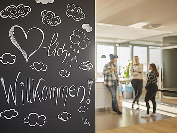 Foto von der FRÖBEL Akademie in Köln. Auf einer Wand mit schwarzer Tafelfarbe steht mit weißer Kreide "Herzlich willkommen!"