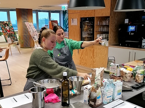 Zwei Frauen stehen nebeneinander an einer Küchenzeile und bereiten Zutaten vor. Die Frau links trägt einen grünen Pullover, die Frau rechts trägt eine grüne Kochjacke mit Fröbel-Logo und eine graue Schürze. Auf der Arbeitsfläche liegen verschiedene Lebensmittel und Kochutensilien. Im Hintergrund sind farbenfrohe Plakate und große Fenster mit Blick auf Köln zu sehen.