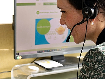 Foto einer Frau, die mit einem Headset am Schreibtisch sitzt und an einem Video-Meeting teilnimmt.