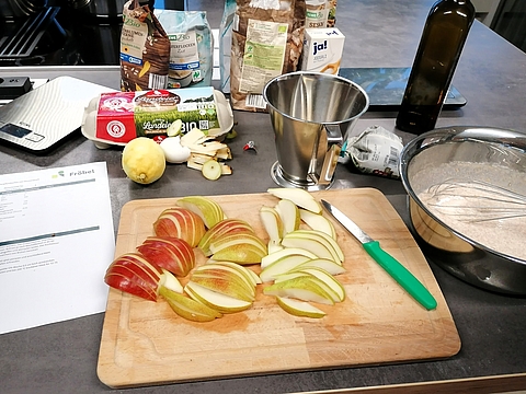 Auf einer Küchenarbeitsfläche liegen vorbereitete Zutaten für ein Rezept: Auf einem Holzbrett sind fein geschnittene Apfel- und Birnenspalten zu sehen, daneben ein Messer mit grünem Griff. Im Hintergrund stehen verschiedene Zutaten wie Mehl, Eier, Öl, Zitronen und Milch, daneben eine Rührschüssel mit Teig und ein Rezeptblatt mit dem Fröbel-Logo.