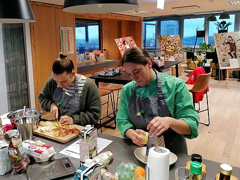 Zwei Frauen stehen an einer Küchenzeile und bereiten frische Zutaten zu. Die Frau links schneidet Äpfel auf einem Schneidebrett, die Frau rechts – in grüner Kochjacke und grauer Schürze mit Fröbel-Logo – reibt eine Zutat. Auf der Arbeitsfläche liegen verschiedene Lebensmittel und Kochutensilien. Im Hintergrund sind bunte Plakate und große Fenster mit Blick über Köln zu sehen.