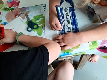 Zwei Kinder sitzen an einem Tisch und arbeiten gemeinsam an einem Bastelprojekt. Sie malen Verkehrsschilder aus. Das Kind rechts im Bild hält mit einer Hand das Papier fest und koloriert mit einem blauen Wachsmalstift ein Verkehrsschild mit der Aufschrift „Einbahnstraße“. Das Schild ist rechteckig und mit einem Pfeil nach rechts versehen. Das andere Kind, links im Bild, koloriert mit einem roten Stift ein weiteres Verkehrsschild, das nur teilweise sichtbar ist. Beide Kinder sind konzentriert bei der Arbeit. Auf dem Tisch liegen außerdem eine Kinderschere mit grünen Griffen und weitere Bastelmaterialien. Der Tisch ist mit einer bunten, blumig gemusterten Folie bedeckt. Im Hintergrund sieht man einen Kinderfuß, der barfuß auf dem Boden steht. Die Szene vermittelt eine kreative Lern- und Spielsituation