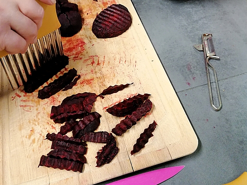 Eine Hand hält einen gezackten Wellenschneider und schneidet rote Bete in gleichmäßige Stücke. Auf dem Holzbrett liegen bereits geschnittene Stücke, daneben ein Sparschäler und ein pinkes Messer.