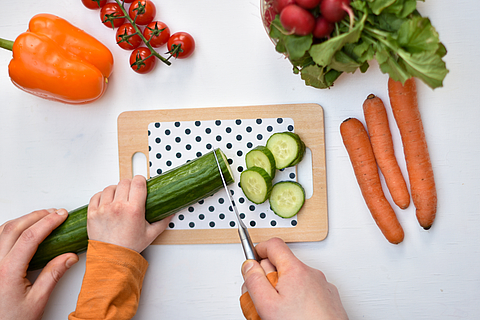 Kinderhände schneiden auf einem Schneidebrett sorgfältig Gemüse wie eine Gurke.